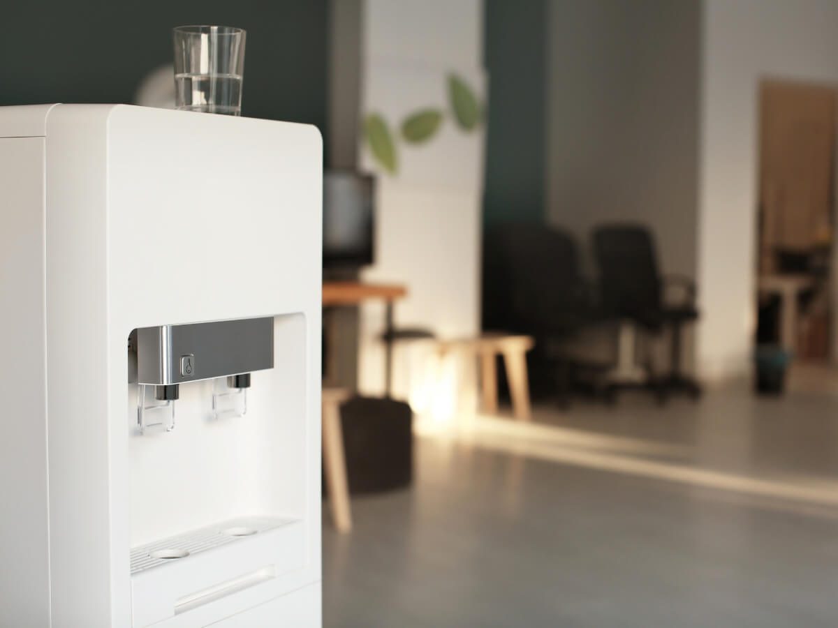 Image of a bottleless water cooler with a glass of water on top, set against a background of a workplace, emphasizing the integration of sustainable hydration solutions in office environments.